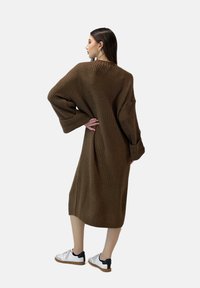 Bruine gebreide jurk met oversized mouwen en een losse pasvorm, voorzien van een gestructureerd patroon. Model combineerde het met witte sneakers.