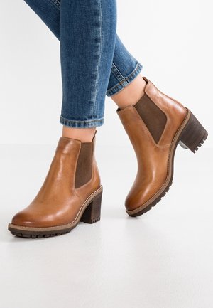 Person iført brune læderankelstøvler med blokhæl og blå jeans mod en hvid baggrund.