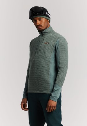 Man die een groene textuurige trui met rits draagt, bijpassende muts met zwarte skibril en donkere broek, staand tegen een effen achtergrond.