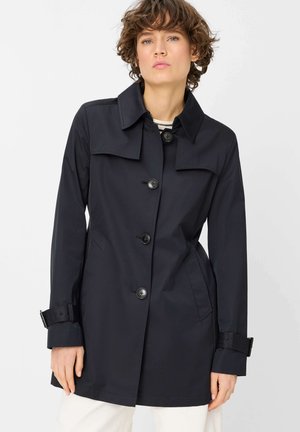 Frau mit kurzen lockigen Haaren, die einen schwarzen Trenchcoat mit Knöpfen und Schnallenriemen an den Ärmeln sowie weiße Hosen vor weißem Hintergrund trägt.