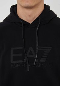 Felpa nera con cappuccio e coulisse, con un logo "EA7" in rilievo e il testo "Emporio Armani" nello stesso colore. Texture morbida.
