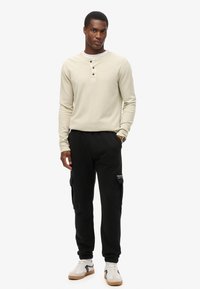 Jasno beżowy longsleeve henley z trzema guzikami, w zestawie z czarnymi spodniami cargo z bocznymi kieszeniami oraz białymi trampkami z brązowymi akcentami.
