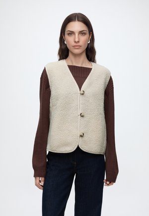 Femme portant un gilet polaire crème boutonné sur un pull marron à manches longues avec un jean foncé, debout devant un fond blanc uni.