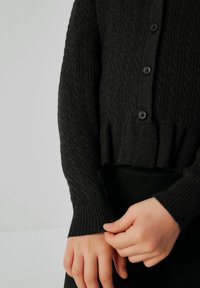 Cardigan nero a coste con un motivo a trecce, dotato di chiusura con bottoni e orlo corto. Le maniche sono aderenti con polsini a coste.