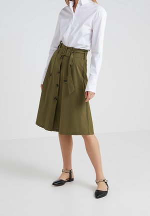 Mujer con camiseta blanca de manga larga, falda hasta la rodilla color verde oliva con botones y cinturón, y bailarinas negras con tiras en el tobillo, de pie frente a un fondo blanco.
