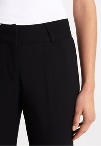 Pantalon noir sur mesure avec une texture lisse, doté d'un devant plat, de poches inclinées et d'une ceinture ajustée. Gros plan sur la jambe du pantalon et la main.
