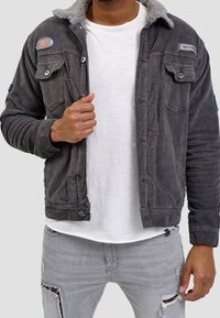 Grijze corduroy jas met een zachte kraag, knoopsluiting aan de voorkant, twee borstzakken en patches op de linkerschouder en de bovenkant van de borst. Gedragen over een wit shirt.