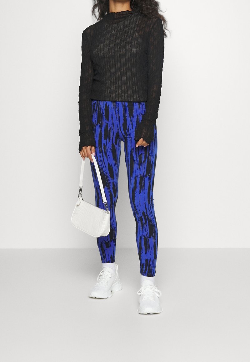 Haut noir à manches longues côtelé, leggings à motifs bleus et noirs, baskets blanches et un petit sac blanc avec une bandoulière.