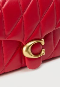 Κόκκινη δερμάτινη τσάντα με καπιτονέ σχέδιο, με χρυσό κούμπωμα σε σχήμα ημισελήνου και διαγώνιο μοτίβο ραφής για υφή και λεπτομέρεια.