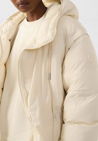 Donsjack in licht crèmekleur, met capuchon, dikke isolatie, ritssluiting en een gestructureerde afwerking. Gladde stof met een aansluitend ontwerp.