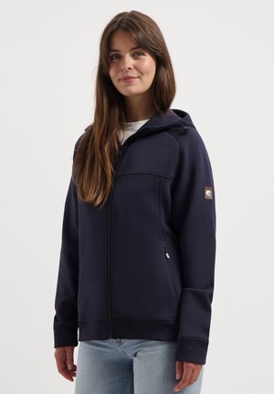 Young woman with long brown hair wearing a navy zip-up hoodie and light blue jeans, standing against a plain white background.