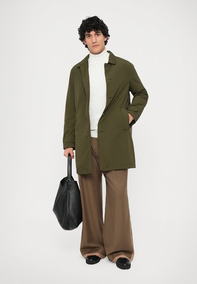 Muškarac s kovrčavom kosom nosi maslinasto zeleni kaput, bijeli turtleneck, široke smeđe hlače, crne cipele, te drži veliku crnu kožnu torbu.