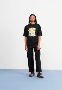 T-shirt nero oversize con un design grafico colorato al centro, abbinato a pantaloni cargo neri e scarpe da ginnastica marroni.