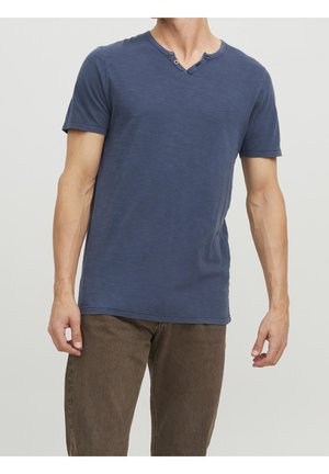 Mann trägt ein dunkelblaues Henley-Shirt mit kurzen Ärmeln und braune Hose, steht vor einem schlichten hellen Hintergrund.