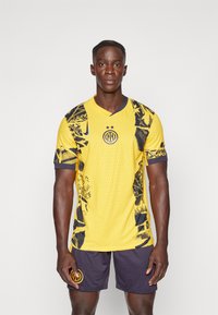 Kollane jalgpallipaita mustade geomeetriliste mustrite ja aktsentidega, millel on V-kaelus ja logo. Kanda tumesiniste lühikeste pükstega.