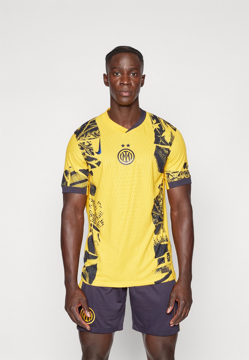 Kollane jalgpallipaita mustade geomeetriliste mustrite ja aktsentidega, millel on V-kaelus ja logo. Kanda tumesiniste lühikeste pükstega.