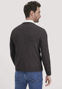 Mann, der seitlich steht und einen dunkelgrauen Pullover über einem weißen Hemd mit Kragen und blauen Jeans trägt, vor einem einfarbigen Hintergrund.