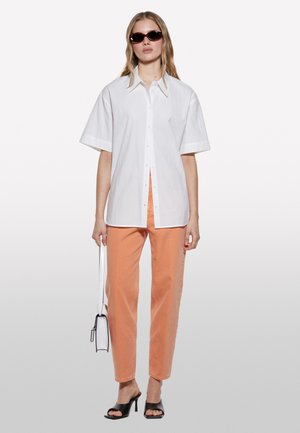 Chemise blanche à manches courtes, associée à un pantalon droit orange clair. Sandales à talons noires et petit sac à main blanc.
