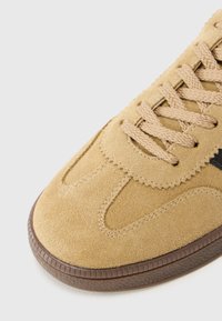 Beige suede sneaker with textured surface, contrasting dark accent, round toe, and patterned rubber sole. Features flat laces and stitched details.