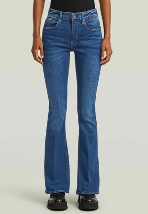 Persona che indossa jeans bootcut blu a vita alta e scarpe platform nere, in piedi davanti a uno sfondo chiaro e uniforme.