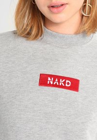 Sudadera gris con un cuello alto, que presenta un parche rectangular rojo con el texto "NAKD" en bordado blanco. Textura suave, ajuste casual.