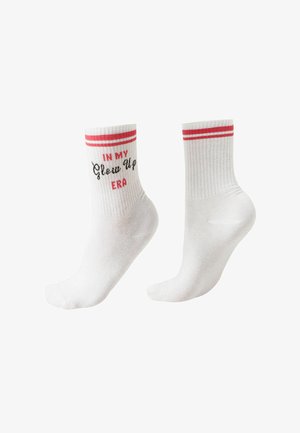 Białe skarpetki za kostkę z czerwonymi paskami na górze; jedna skarpetka ma z boku napis "IN MY Glow Up ERA" w czerwono-czarnych literach.