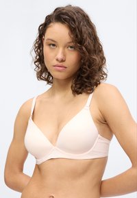 Νεαρή γυναίκα με σγουρά μαλλιά, φοράει ανοιχτό ροζ σουτιέν, κοιτάζοντας ευθεία με ουδέτερη έκφραση, πάνω σε λευκό φόντο.