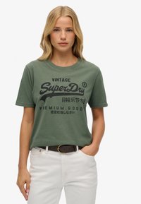 Grünes Baumwoll-T-Shirt mit kurzen Ärmeln, Rundhalsausschnitt und einem Vintage-Design in Schwarz, das den Text und das Logo von "SuperDry" zeigt.