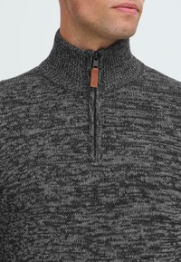 Uomo che indossa un maglione a maglia marled grigio scuro e nero con una breve cerniera e un tab in pelle marrone al collo.
