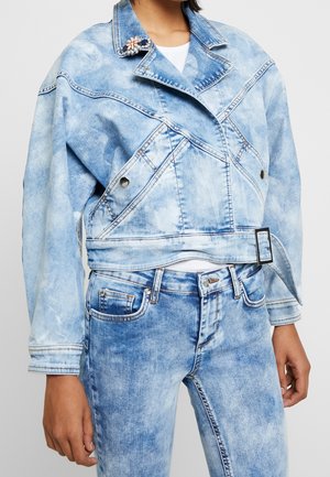 Person trägt eine hellblaue Acid-Wash-Denimjacke mit floralem Broschen-Anstecker und passenden hoch taillierten Jeans, Hände entspannt seitlich.