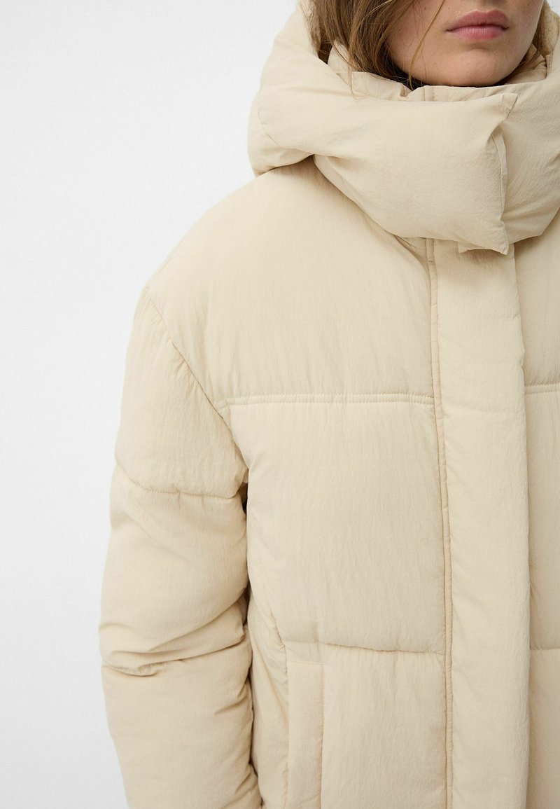 Giubbotto imbottito beige con colletto alto e imbottito, cuciture orizzontali e tasche laterali. Tessuto morbido e testurizzato con una finitura liscia.