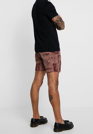 Mann in schwarzem T-Shirt, roten gemusterten Shorts und schwarzen Leder-Loafers, der mit verschränkten Armen vom Kamerablick abgewandt steht.