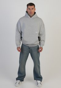 Felpa grigia chiara con tasca frontale e logo discreto, abbinata a jeans blu a vestibilità ampia e sneakers bianche. Tessuto morbido, design rilassato.