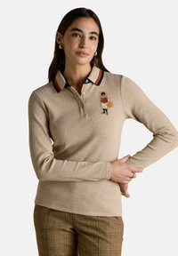 Frau in einem beigefarbenen Langarm-Polo mit Fuchs-Stickerei auf der Brust, gestreiftem Kragen und karierter brauner Hose, steht mit einem angewinkelten Arm.