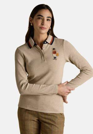 Frau in einem beigefarbenen Langarm-Polo mit Fuchs-Stickerei auf der Brust, gestreiftem Kragen und karierter brauner Hose, steht mit einem angewinkelten Arm.