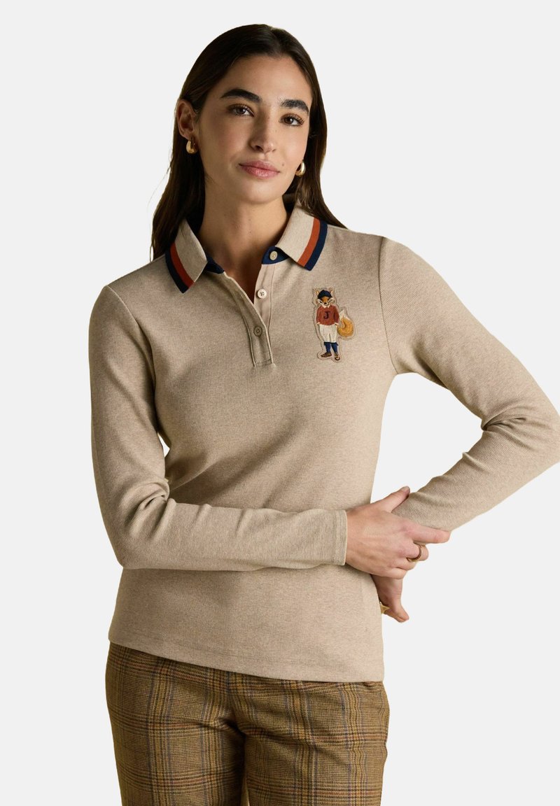 Frau in einem beigefarbenen Langarm-Polo mit Fuchs-Stickerei auf der Brust, gestreiftem Kragen und karierter brauner Hose, steht mit einem angewinkelten Arm.
