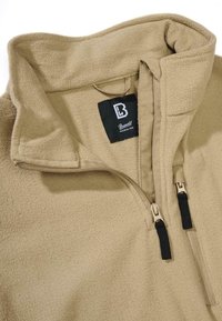 Beige Fleecejacke mit hohem Kragen, halbem Reißverschluss und kontrastierenden schwarzen Reißverschlussgriffen. Etikett ist im Kragen sichtbar. Weiche Textur.