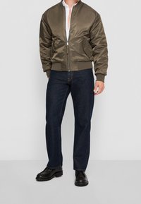 Olijfgroene bomberjack met ritssluiting, geribbelde manchetten en zoom, gecombineerd met donkerblauwe jeans en zwarte schoenen. Gladde stof met een strak design.