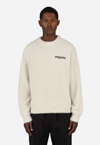 Svetlo bež pulover z mehkobo in puhastim materialom. Ima okroglo ovratnico in dolge rokave. Na prsih je izvezen napis "Pagador" v temni nitki.