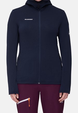 Personne portant un sweat à capuche zippé bleu marine avec le logo "Mammut" sur la poitrine et un pantalon bordeaux avec une poche zippée blanche et un onglet orange.