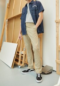 Man wearing beige pants, black and white sneakers, gray t-shirt, and open short-sleeve denim shirt in a workshop setting.
