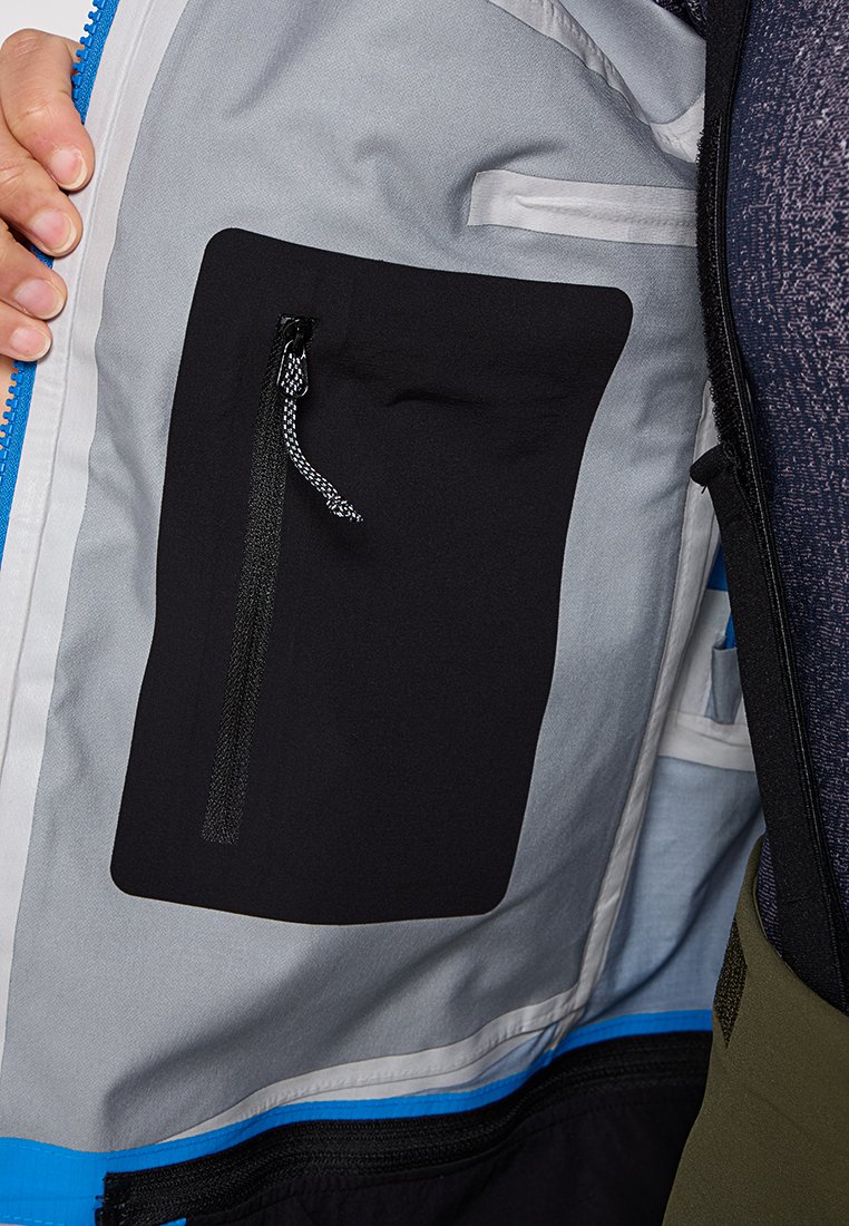 Schwarze Innentasche mit Reißverschluss auf einem hellgrauen Stoff, am Rand von einem blauen Akzent umrandet. Strukturierte Fütterung sichtbar.