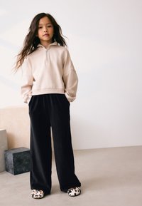 Svijetlosmeđi duks s patentnim zatvaračem, crne hlače širokih nogavica i papuče s uzorkom kravljeg mlijeka. Glatki materijal i opušteni kroj su istaknute značajke.