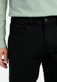 Zwarte denim jeans met een textuurafwerking, voorzien van een klassiek vijf-pocket ontwerp en knoopsluiting, gecombineerd met een lichtgroen shirt met lange mouwen.