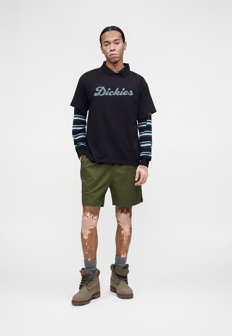 Junger Mann mit Vitiligo, der ein schwarzes Dickies-Polo über gestreiften Langarmshirts, olivgrüne Shorts, graue Socken und braune Stiefel trägt, steht vor weißem Hintergrund.