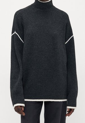 Pull à col roulé gris foncé avec bordure blanche aux poignets, à l'ourlet et des rayures en biais sur le haut des manches, porté avec un pantalon noir.