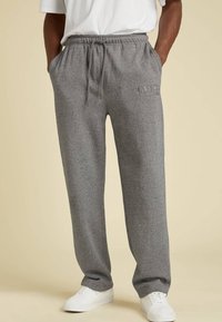 Hombre vistiendo pantalones de chándal grises con cordón y el logo de "GUESS Originals", zapatillas blancas, con las manos en los bolsillos, de pie sobre un fondo beige.