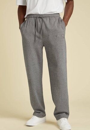 Homme portant un pantalon de survêtement gris avec le logo "GUESS Originals" et des baskets blanches, les mains dans les poches, se tenant sur un fond beige.