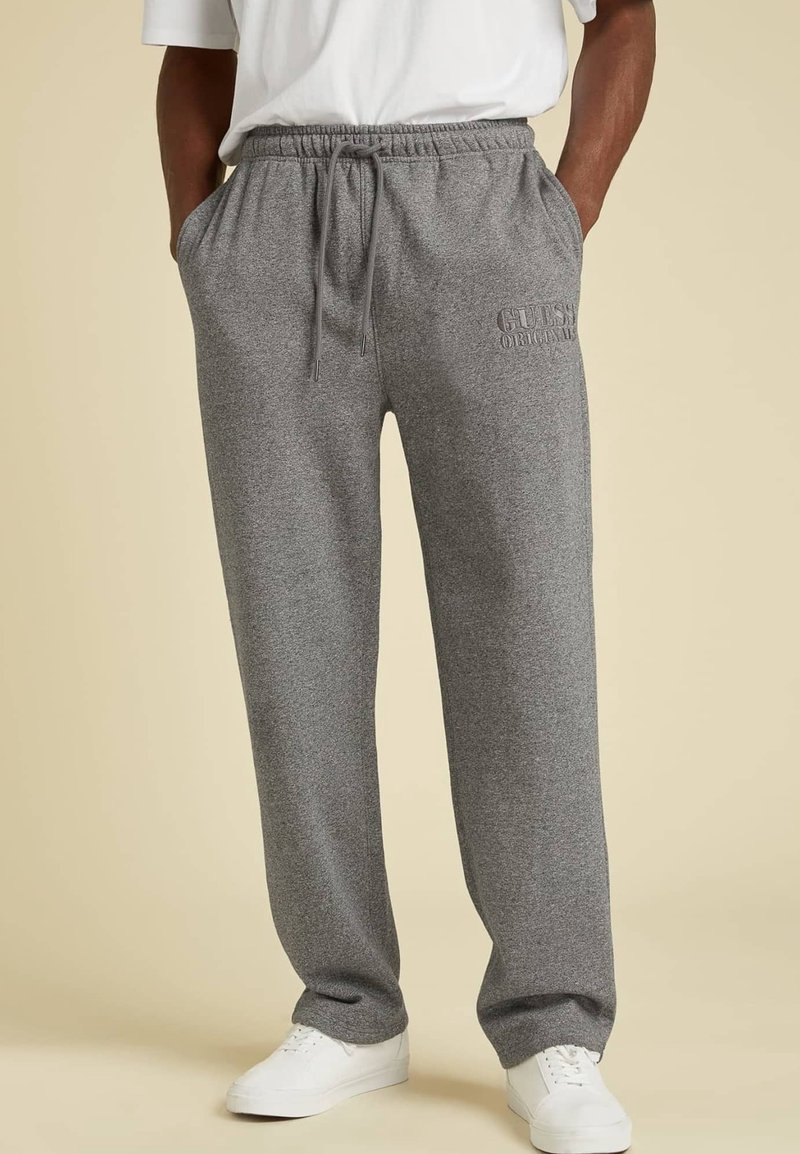 Hombre vistiendo pantalones de chándal grises con cordón y el logo de "GUESS Originals", zapatillas blancas, con las manos en los bolsillos, de pie sobre un fondo beige.