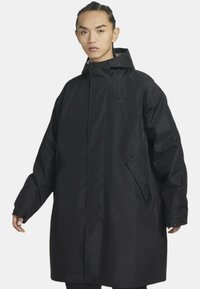 Cappotto impermeabile nero con colletto alto, maniche lunghe e vestibilità morbida. Presenta una zip frontale, tasche laterali e un sottile logo decorativo.